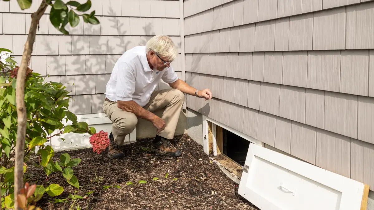Inspector kneeling at the crawl space vent of a house with gray siding